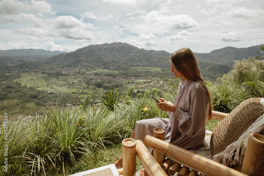 Young girl with a smartphone sits on a bamboo chair with a beautiful view of the mountains or the volcano. Bamboo house in Bali.