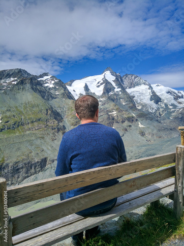 Grossglockner, Austria - August 17, 2019: Grossglockner high alpine road against the blue sky. Man sits on a wooden bench and admires the peaks of mountain peaks. Vertical