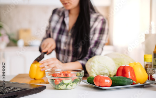 A cheerful young woman cooks in the kitchen with an online audience. The girl prepares a salad  slices a cucumber during an online broadcast. online cooking. selective focus