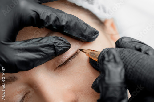 Master makes permanent tattoo makeup on eyelashes of eyes of young woman in beauty salon