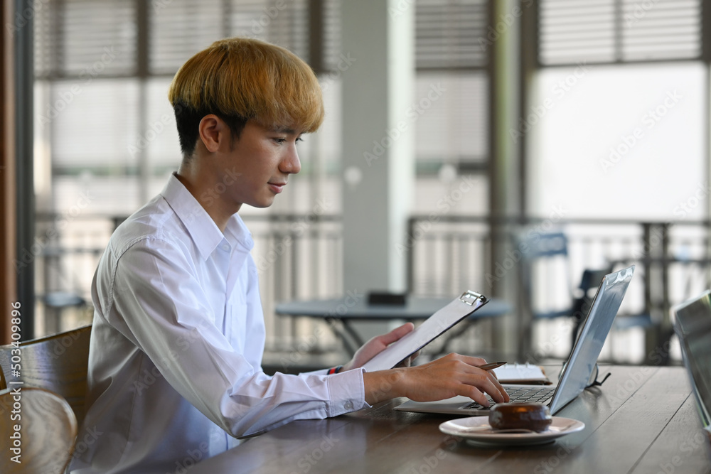 Side view asian male employee holding document and using laptop computer at office desk.