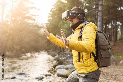 backpacker man in the forest with virtual reality headset looking directly and trying to touch something with his hand - metaverse concept -
