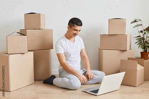 Indoor shot of brunette man sitting on floor with laptop before to leave apartment, happy Caucasian guy among cardboard boxes ordering delivery. Distance work from any place and freelance.