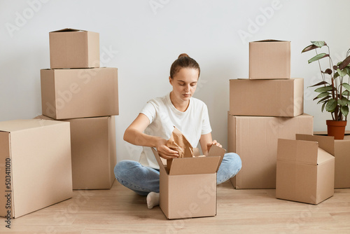 Indoor shot of adorable delighted female in white t-shirt packing cardboard box, woman packing things to move among carton parcels, relocation and moving to new house. © sementsova321