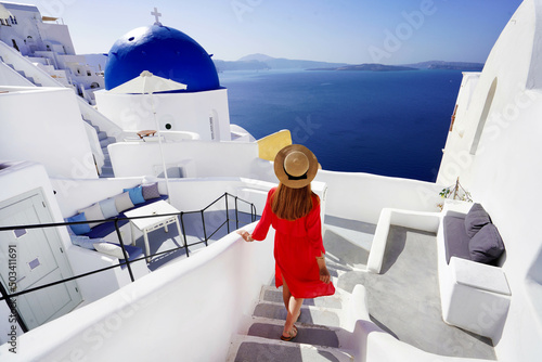 Beautiful girl in red dress and hat comes down the stairs to her resort enjoying a spectacular view of the Caldera in Oia village, Santorini, Greece