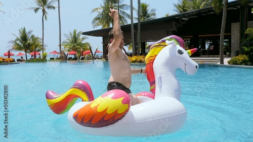 Funny video young man falling in pool in luxury hotel, resort. Happy man have fun ride dance on giant inflatable unicorn in swimming pool, splashing, enjoy holidays or vacations on sunny day in summer photo