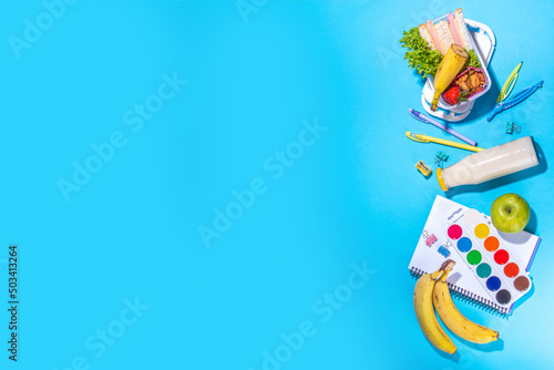 Healthy school lunch box: sandwich, vegetables, fruit, nuts and yogurt with school kids supplies, accessories and backpack on high-colored blue background flatlay copy space