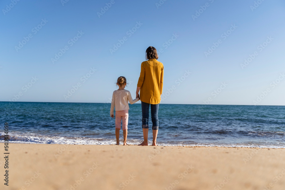 Woman with daughter standing on shore looking at horizon Woman with daughter standing on shore looking at horizon