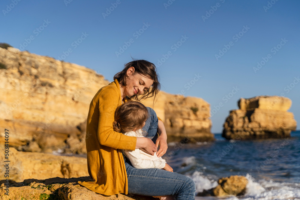 Smiling woman with daughter sitting on rock at beach Smiling woman with daughter sitting on rock at beach