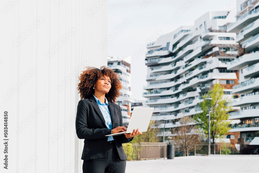 Thoughtful businesswoman holding laptop standing by white wall on sunny day Thoughtful businesswoman holding laptop standing by white wall on sunny day