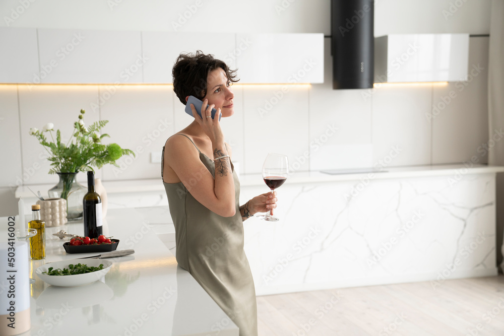 Woman holding wineglass talking on mobile phone standing in kitchen Woman holding wineglass talking on mobile phone standing in kitchen