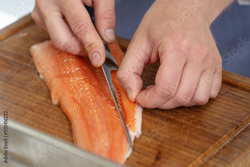 Hands of a cook remove the belly flap with bones of a fresh raw char fish with a filleting knife on a cutting board, selected focus photo