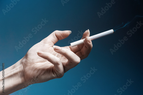 World No Tabacco day. A pale, crooked female hand, close-up, holding a smoking cigarette. Dark blue background. The concept of nicotine addiction