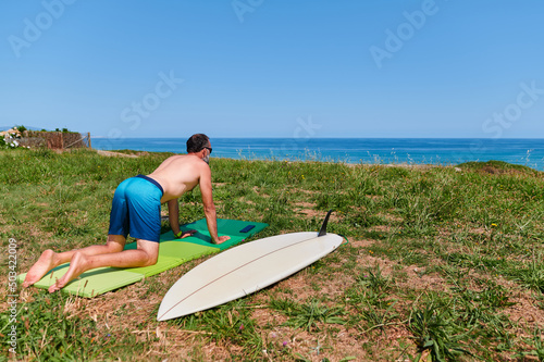 Back view of mature male surfer kneeing on mat on seashore and stretching lower back while preparing for surfing on sunny day in summer © Enadan