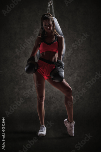 Fototapeta Naklejka Na Ścianę i Meble -  Athletic woman in red shorts and top posing near the bag. Boxing and mixed martial arts concept
