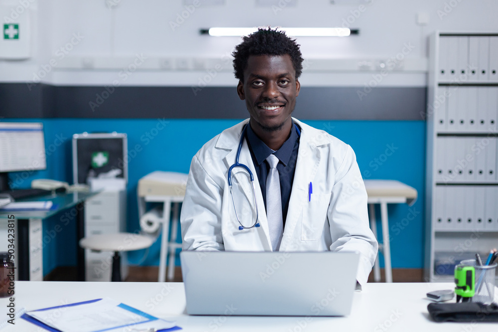 Smiling african american healthcare clinic specialist sitting at desk while using work laptop. Hospital doctor wearing white coat uniform sitting at desk in workspace while using modern computer Smiling african american healthcare clinic specialist sitting at desk while using work laptop. Hospital doctor wearing white coat uniform sitting at desk in workspace while using modern computer