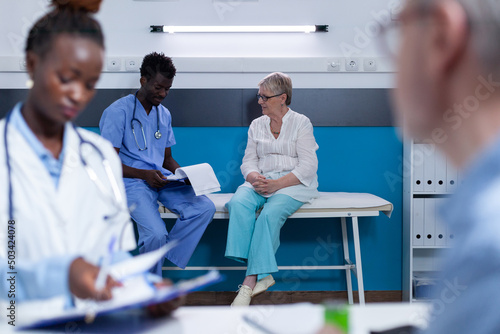 Hospital nurse conversating with sick retired woman about illness diagnostic and health risks. Clinic staff analyzing disease results and medical file for treatment schedule and medical expertise.