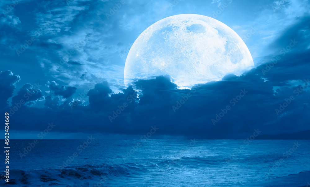 Night sky with full bright moon in the clouds, blue sea in the foreground 