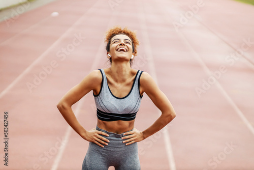 Fototapeta Naklejka Na Ścianę i Meble -  A happy runner stands in the stadium with her hands on hips and laughing. She is so happy she accomplished her goal and moved boundaries. A runner resting and laughing.