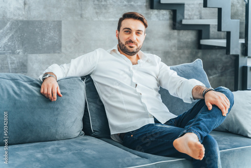 Brown-haired handsome, barefoot confident and smiling bearded businessman looking at camera, sit on blue couch at home.