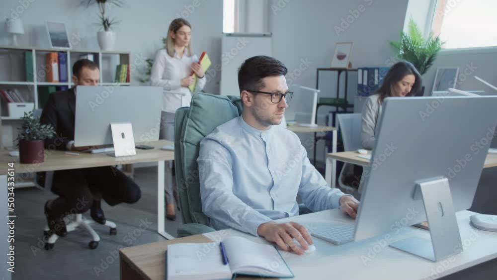 Young attractive man working on computers i talking with colleagues in the office. In the background businesspeople work. Slow-motion Young attractive man working on computers i talking with colleagues in the office. In the background businesspeople work. Slow-motion