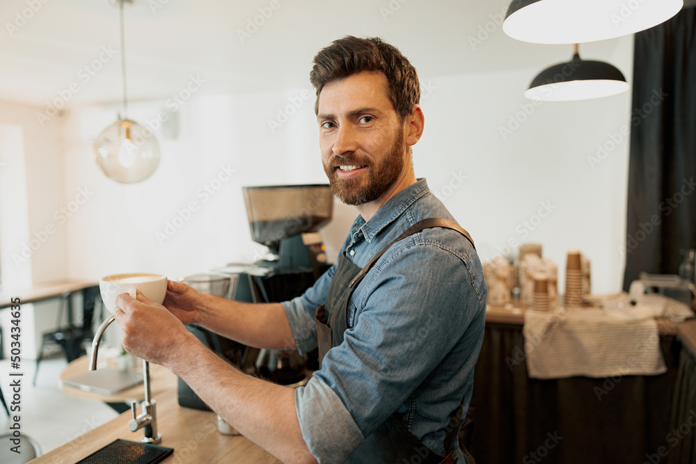 Handsome barista holding cup of coffee and look at the camera standing behind a counter Handsome barista holding cup of coffee and look at the camera standing behind a counter
