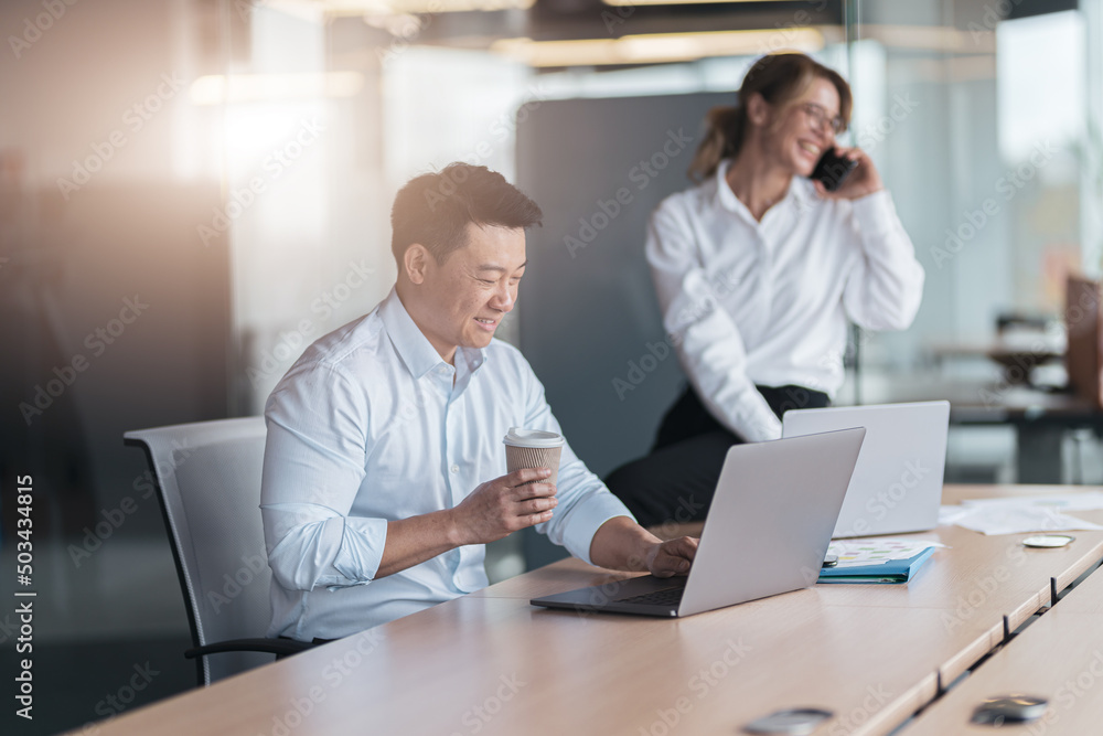 Asian businessman drink coffee and working on laptop with colleague talking on phone in background Asian businessman drink coffee and working on laptop with colleague talking on phone in background