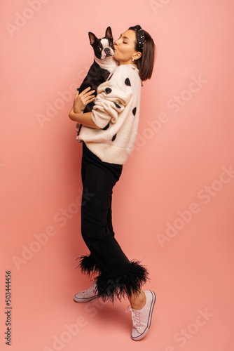 Full length young caucasian woman kissing her black and white dog while standing on pink background. Brunette with bob haircut wears sweater and pants. True friendship, wonderful happy moments photo