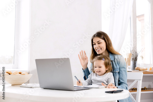 Beautiful mother and little baby works from home, she sits in the kitchen and using laptop for remote work, her cute little girl sits nearby on a chair and waves hello to the family.
