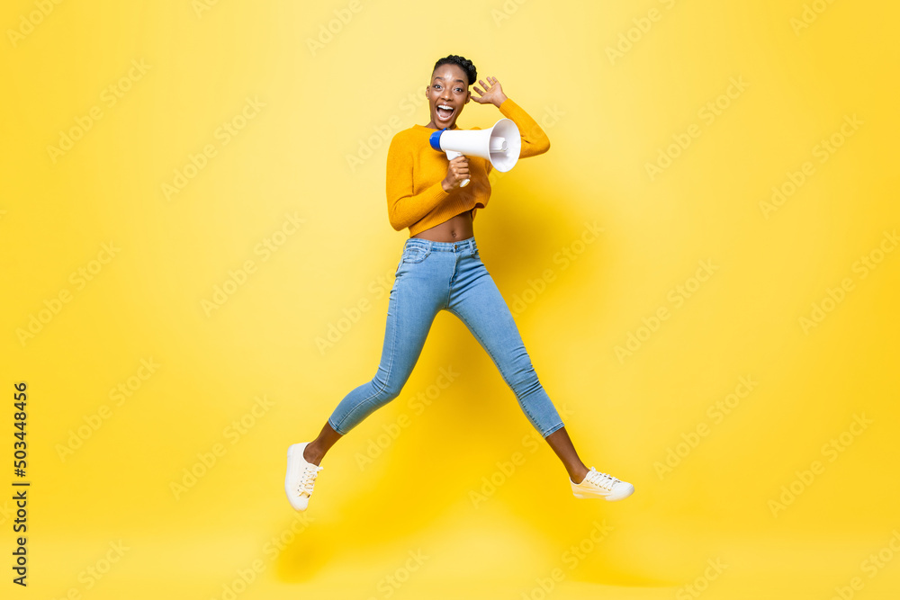 Portrait of cheerful young African American woman holding megaphone jumping in isolated yellow background