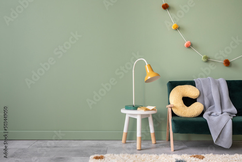 Creative composition of stylish and cozy child room interior design with green wall with bottle greeen sofa, plush pilow, bright carpet, grey plaid and stool with accessories. Copy space. Template.... photo