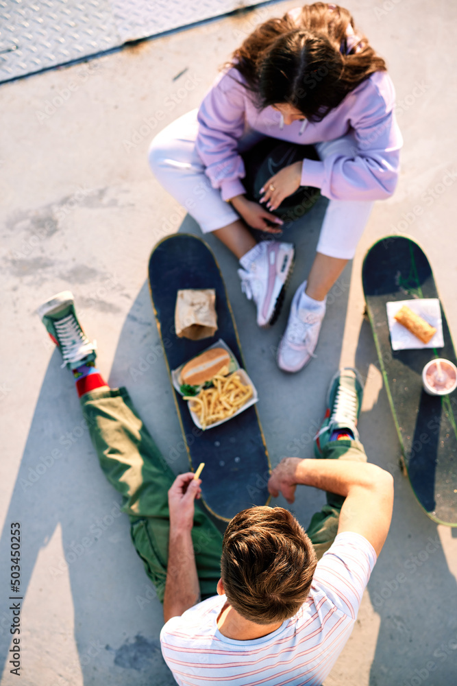 © chika_milan - Top view on teenagers eating fast food on their skateboards. Selective focus on a teenage boy's head who sits with his female friend. © chika_milan - Top view on teenagers eating fast food on their skateboards. Selective focus on a teenage boy's head who sits with his female friend.