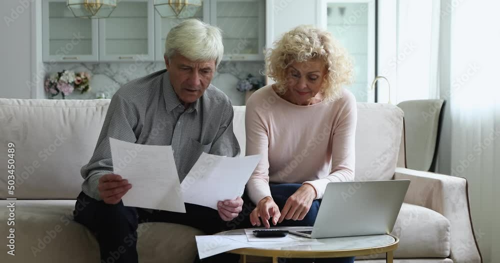 Spouses busy paying bills at home using computer. Serious mature wife and husband sit on sofa managing family budget looking focused, calculate domestic bills, make electronic payment via e-bank app Spouses busy paying bills at home using computer. Serious mature wife and husband sit on sofa managing family budget looking focused, calculate domestic bills, make electronic payment via e-bank app