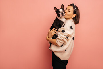 Beautiful young caucasian woman kissing her cute dog on pink background with space for text. Brunette girl in white sweater shares love with pet. Showing affection concept