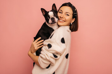 Beautiful young caucasian woman hugging her pet boston terrier breed on pink background. Brunette presses dog to while holding it in her arms. Animal love concept