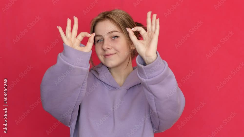 Caucasian smiling young woman attractive in sportswear 20-21 years old shows fingers gesture ok. Studio shot on a red background. Girl shows a sign of consent with her hands, everything is fine Caucasian smiling young woman attractive in sportswear 20-21 years old shows fingers gesture ok. Studio shot on a red background. Girl shows a sign of consent with her hands, everything is fine
