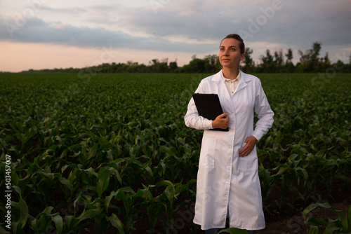 Young woman agronomist in white coat checks growth of corn in field. Farmer takes notes on tablet. agro business concept