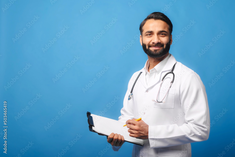 Indian doctor in white medical gown with stethoscope on shoulders taking notes standing with clipboard isolated on blue, physician therapist writing down treatment plan, writes a prescription Indian doctor in white medical gown with stethoscope on shoulders taking notes standing with clipboard isolated on blue, physician therapist writing down treatment plan, writes a prescription