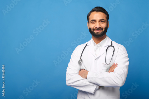 Smiling Indian male doctor pediatric, physical, therapist wearing white medical gown with stethoscope on shoulders stands with arms crossed isolated on blue, copy space. Healthcare and medicine