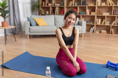 Portrait of young asian lady sitting on fitness mat, taking break from home workout and smiling at camera