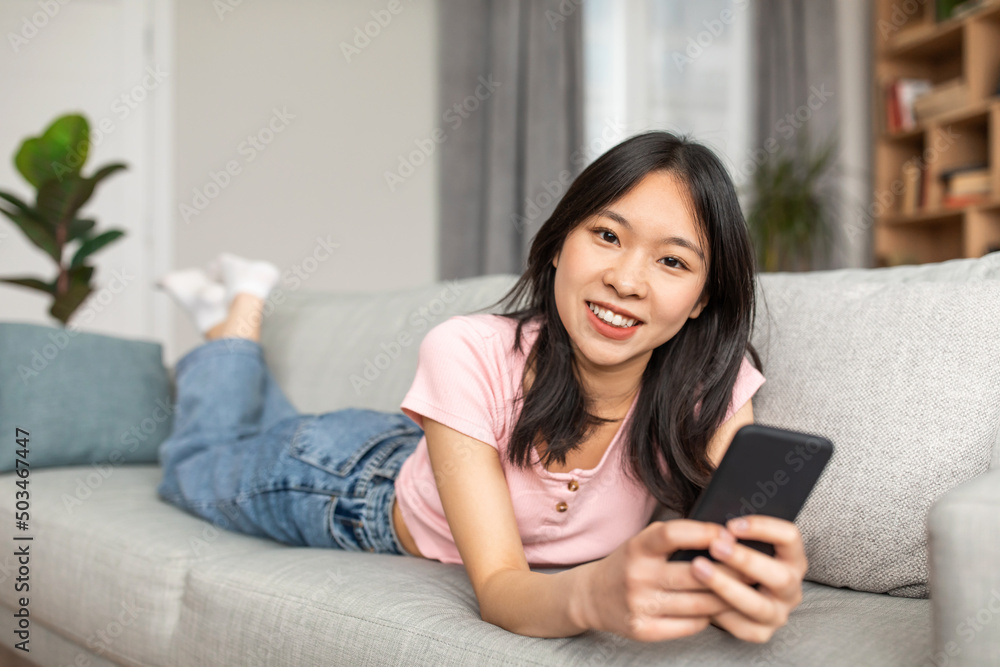 Portrait of positive asian lady using smartphone to work or study, lying on sofa at home and smiling at camera Portrait of positive asian lady using smartphone to work or study, lying on sofa at home and smiling at camera