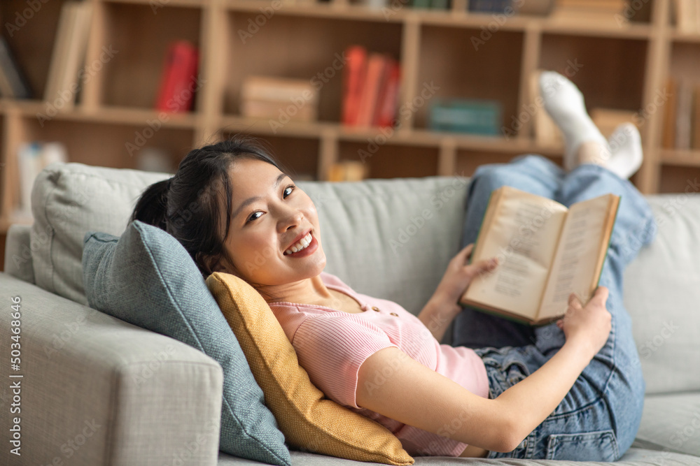 Obraz premium Cheerful asian lady resting on couch, reading book while spending time at home on weekend, smiling at camera