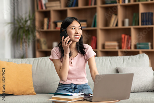 Positive korean lady having phone conversation while sitting on couch at home and using laptop, copy space