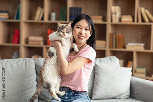 Pretty korean lady posing with her beautiful cat, sitting on sofa in living room, spending time with lovely pet