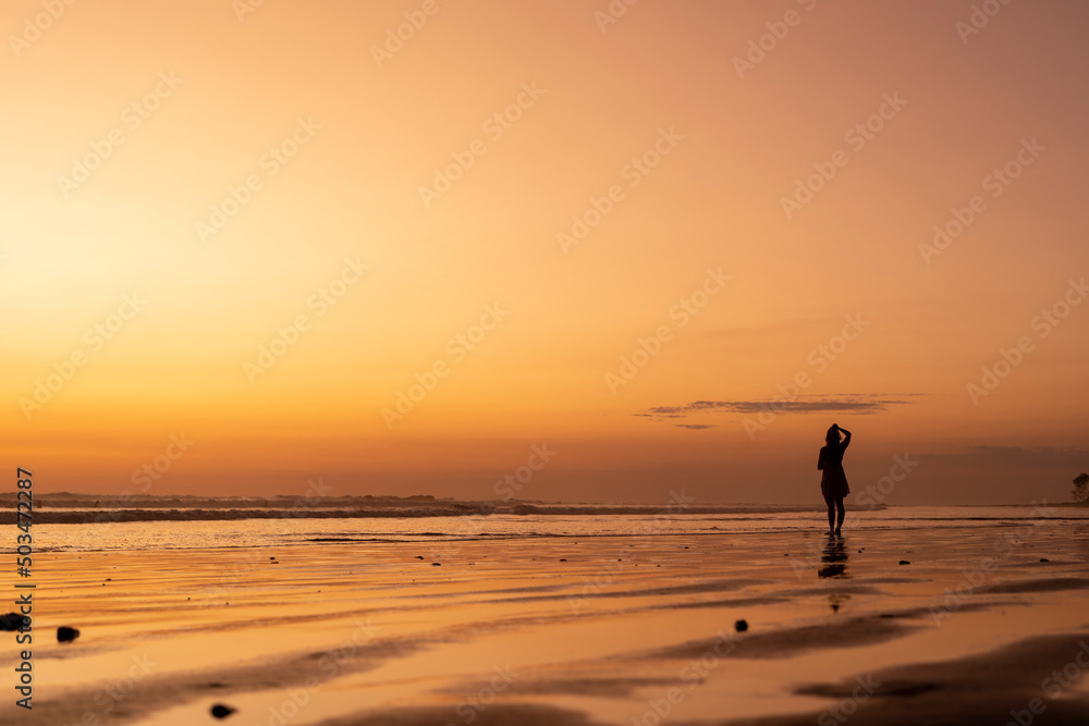 Woman enjoying life on the beach at sunset. Silhouette of young woman standing feeling free and watching golden hour beautiful colors