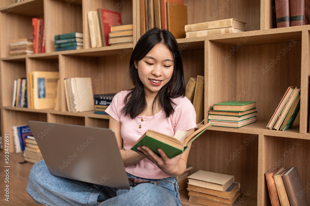 Young korean lady reading book, sitting on floor with laptop pc and study materials at home, copy space Young korean lady reading book, sitting on floor with laptop pc and study materials at home, copy space
