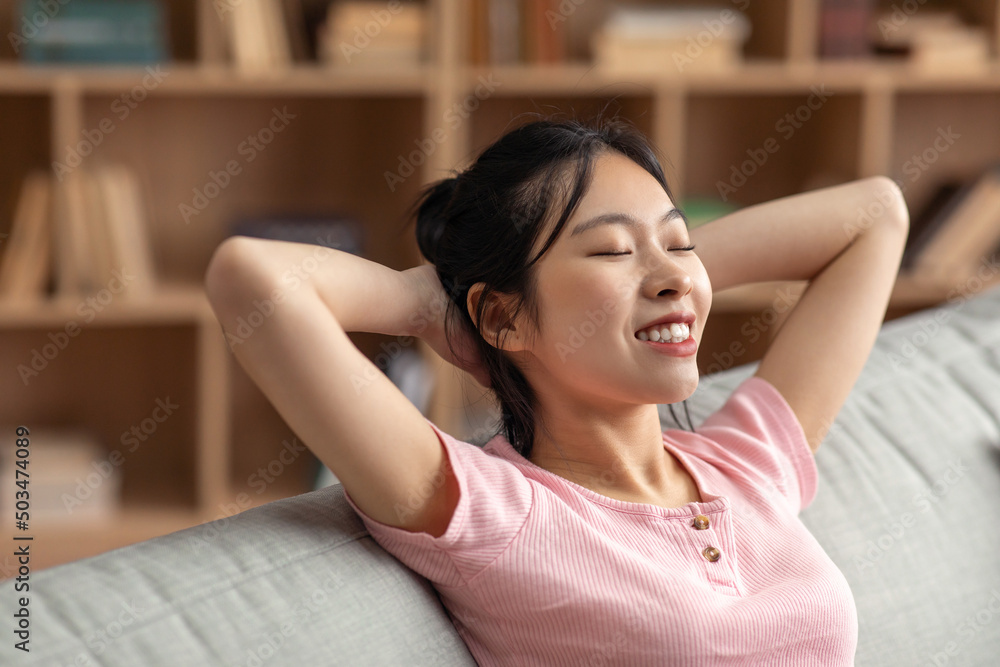 Rest and relax in your own home. Happy asian lady with closed eyes threw hands behind head sitting on sofa Rest and relax in your own home. Happy asian lady with closed eyes threw hands behind head sitting on sofa