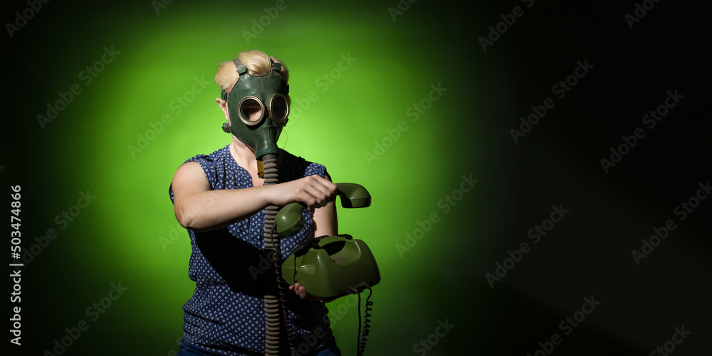 A woman in a gas mask hangs up a retro phone on a dark dramatic background with copy space, hard light.