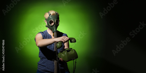A woman in a gas mask hangs up a retro phone on a dark dramatic background with copy space, hard light. © Sergey