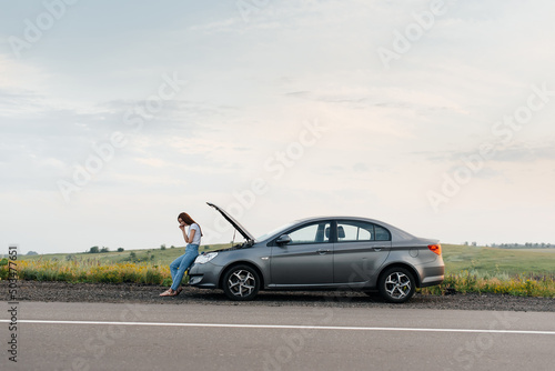 Fototapeta Naklejka Na Ścianę i Meble -  A young girl stands near a broken car in the middle of the highway during sunset and tries to call for help on the phone and start the car. Waiting for help. Car service. Car breakdown on the road.